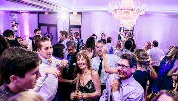 People are dancing and socializing at a party with a chandelier and purple lighting in the background.