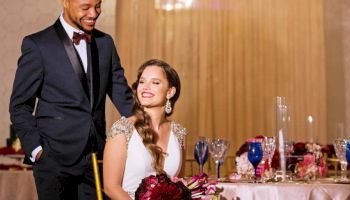 A couple in formal attire at an elegant event under a chandelier, with a decorated table setting and the woman holding flowers.