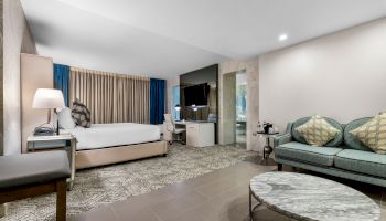 A modern hotel room with a bed, sofa, coffee table, TV, and desk. The decor is sleek, with neutral tones and a mix of textures.