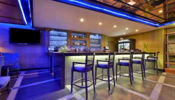 The image shows a modern bar area with high chairs, a backlit bar counter, shelves of glasses and bottles, blue ambient lighting, and a flat-screen TV.