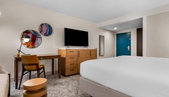 The image shows a modern hotel room with a bed, desk, chair, TV, and abstract wall art, featuring neutral tones and a blue door.