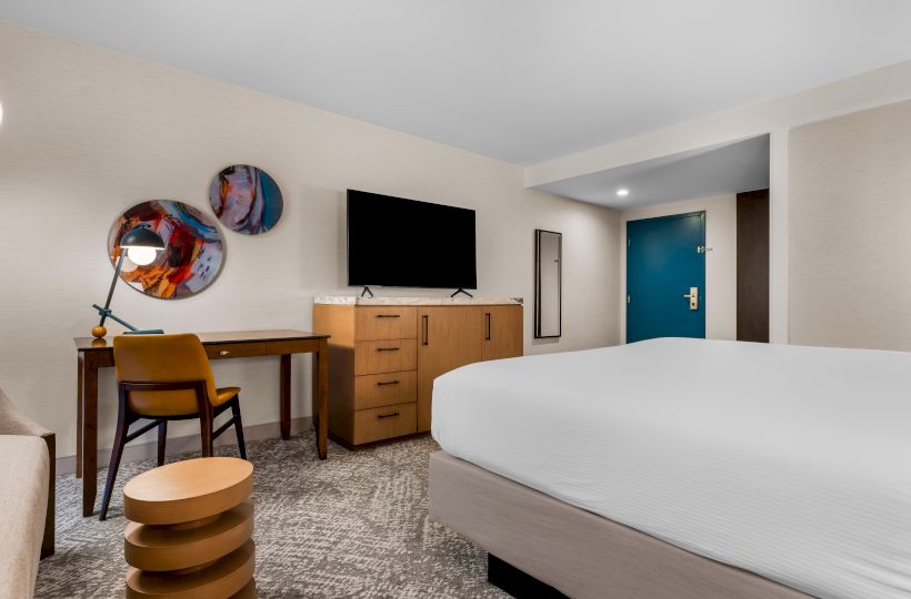 The image shows a modern hotel room with a bed, desk, chair, TV, and abstract wall art, featuring neutral tones and a blue door.