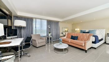 A modern hotel room with a bed, sofa, and armchair, complemented by a desk, lamp, and coffee table in a spacious, well-lit setting.