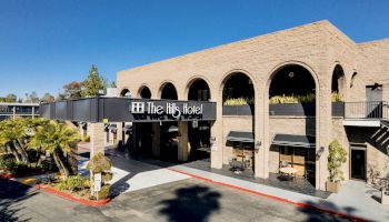 The image shows The Hills Hotel, a beige brick building with large arches and greenery, situated on a sunny day with clear blue skies.