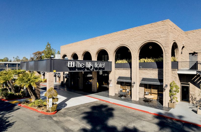 The image shows The Hills Hotel, a beige brick building with large arches and greenery, situated on a sunny day with clear blue skies.