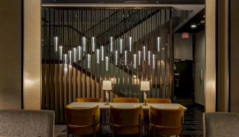 A modern lounge with a long wall of illuminated vertical metal pipes, two tables, chairs, and an &ldquo;EXIT&rdquo; sign down the hallway.