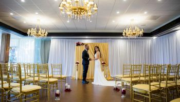 A couple stands in a decorated indoor space, likely a wedding venue, surrounded by chairs, candles, chandeliers, and draped curtains.