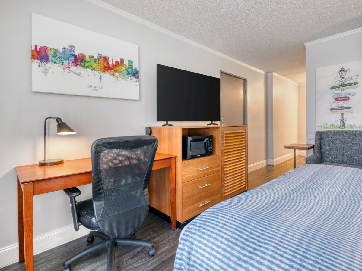 A small office-bedroom with a desk and chair, a TV on a cabinet, a microwave, and colorful city/wizard wall art.