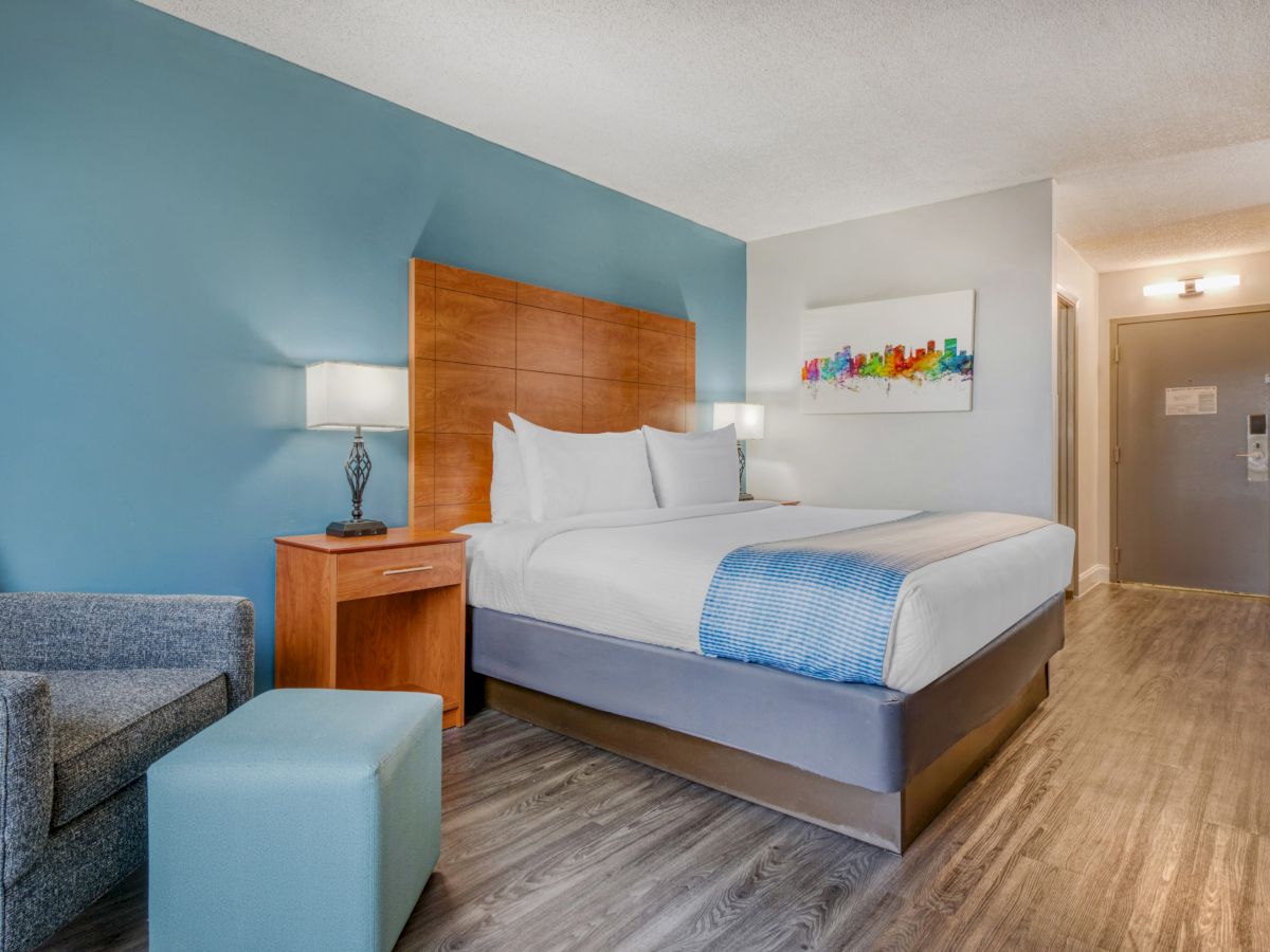 A modern hotel bedroom with a large bed, wood headboard, bedside lamps, seating, and colorful wall art.