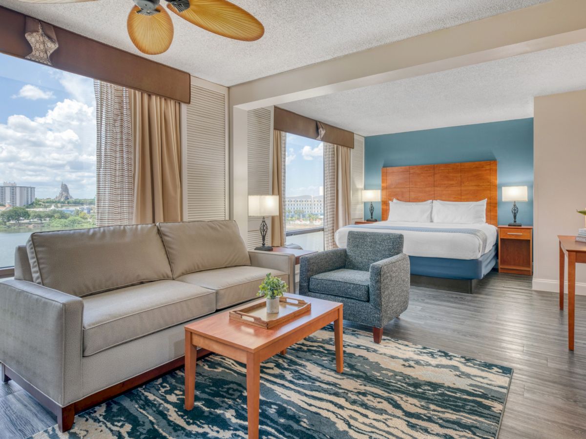 A bright hotel bedroom suite with a sofa, coffee table, bed, bedside lamps, and large windows with curtains and blinds.