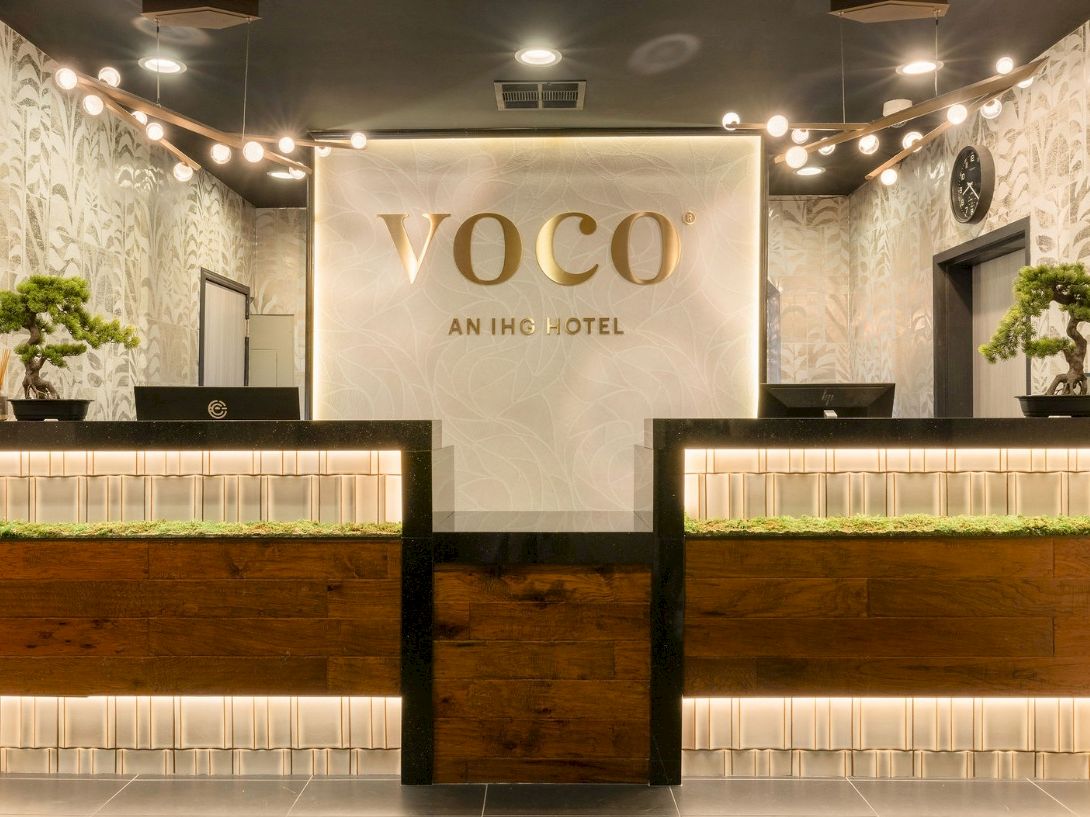 A VOCO hotel reception desk with warm lighting, decorative bonsai trees, and a check-in counter area.