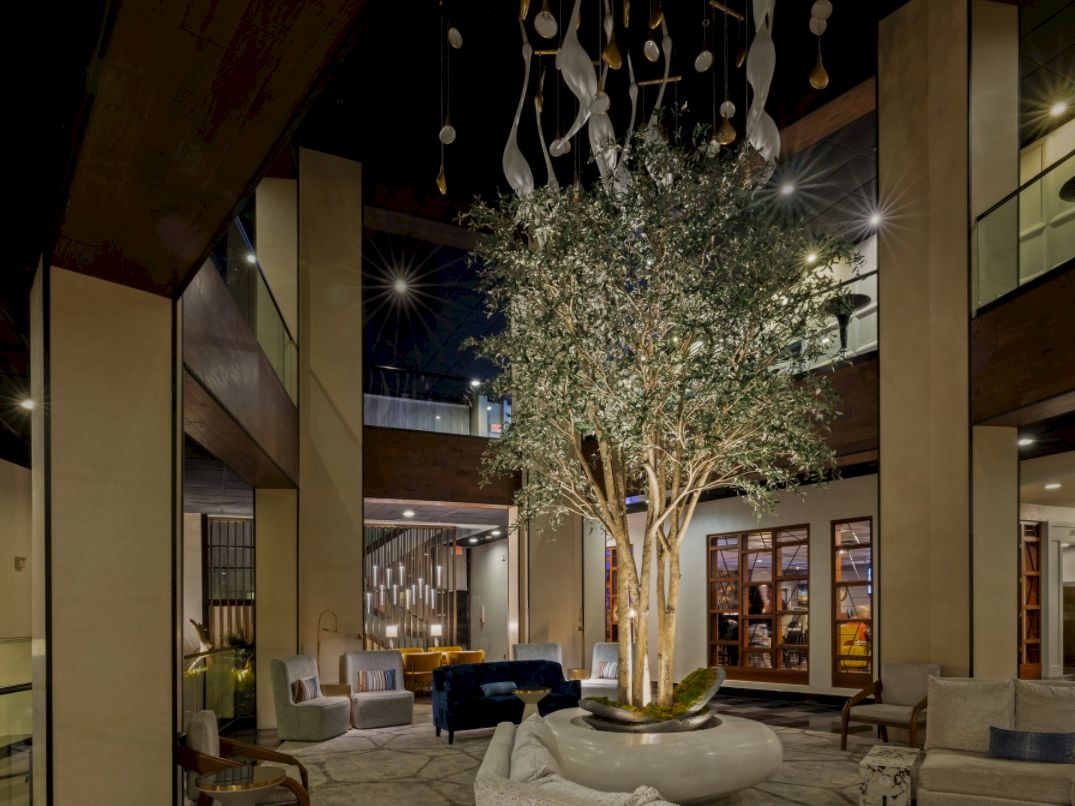 An upscale hotel lobby with a central indoor tree, sofas, pendant lights, and warm marble-and-wood walls.