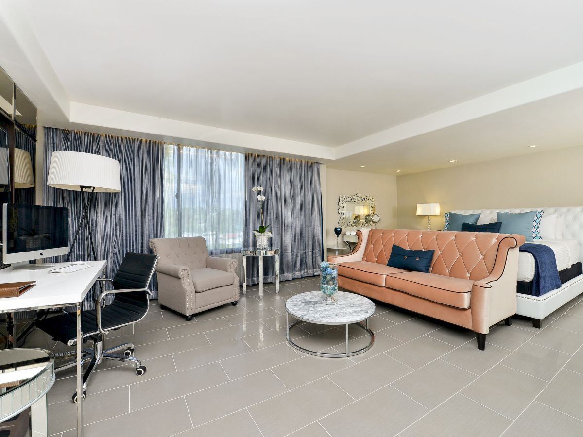 A modern living space with a sofa, armchair, bed, desk, and lamp, decorated in neutral and blue tones, with large windows letting in natural light.