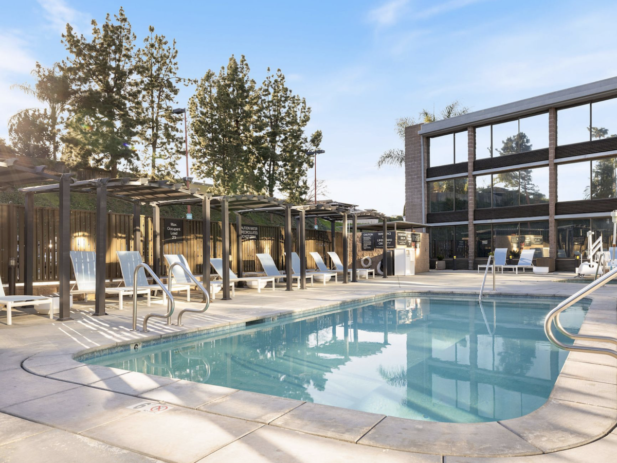 Outdoor hotel pool area with clear blue water, white lounge chairs under pergolas, and a modern brick building under a bright sky.