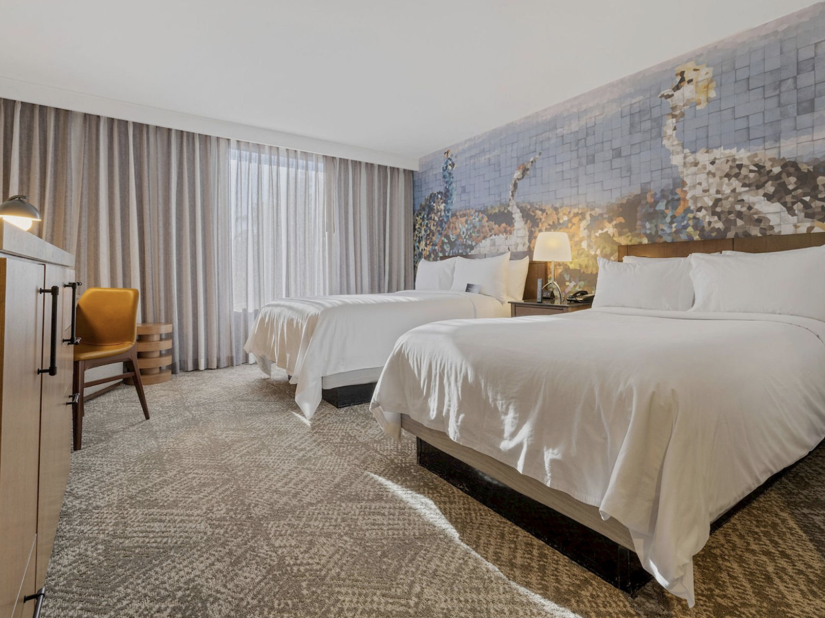 A modern hotel room with two made beds, bedside lamps, sheer curtains, and a large mosaic wall artwork.