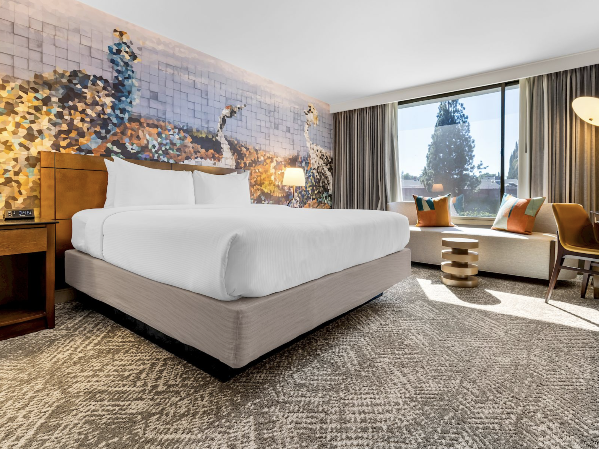 A modern hotel room with a large white bed, colorful wall art, warm lamps, and a view through the window.