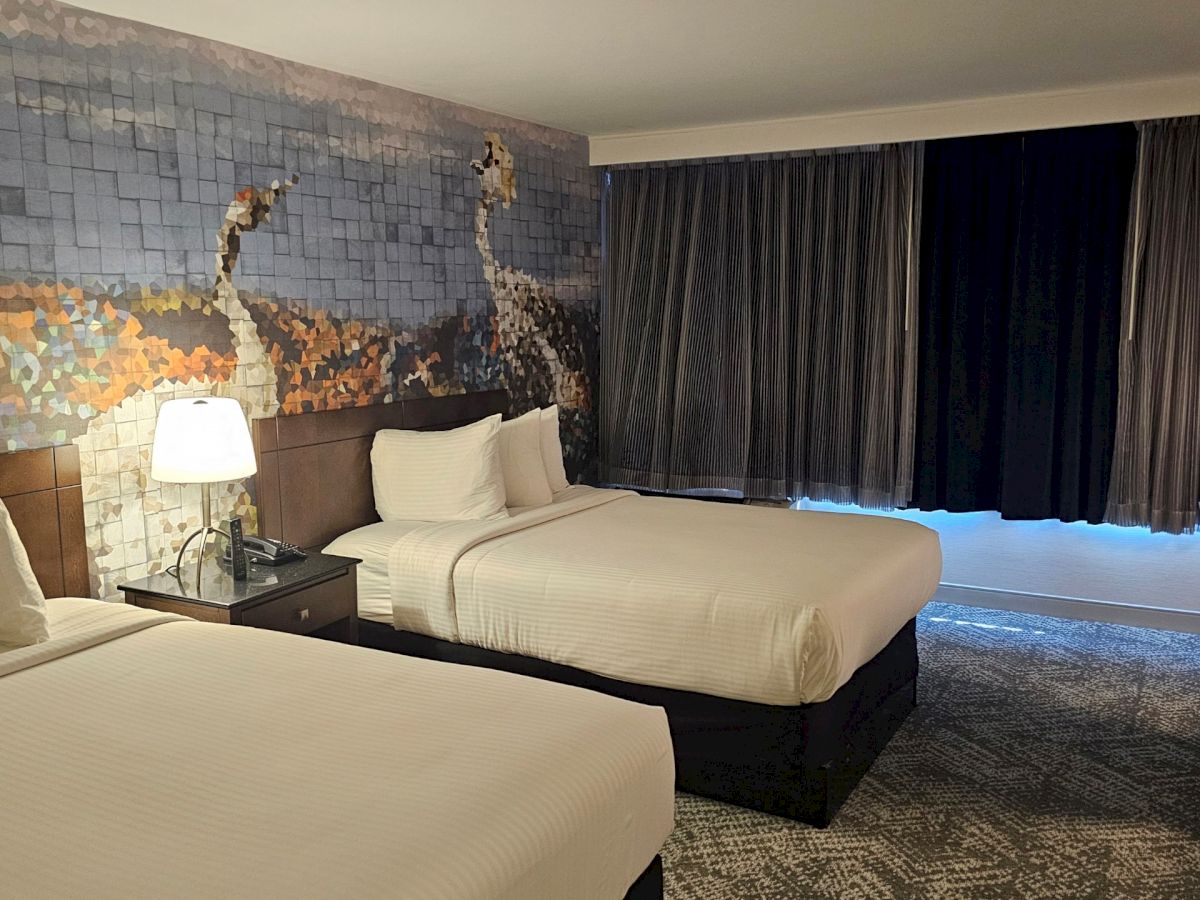 The image shows a hotel room with two beds, a nightstand with a lamp, and a decorative wall mural. The room is softly lit.