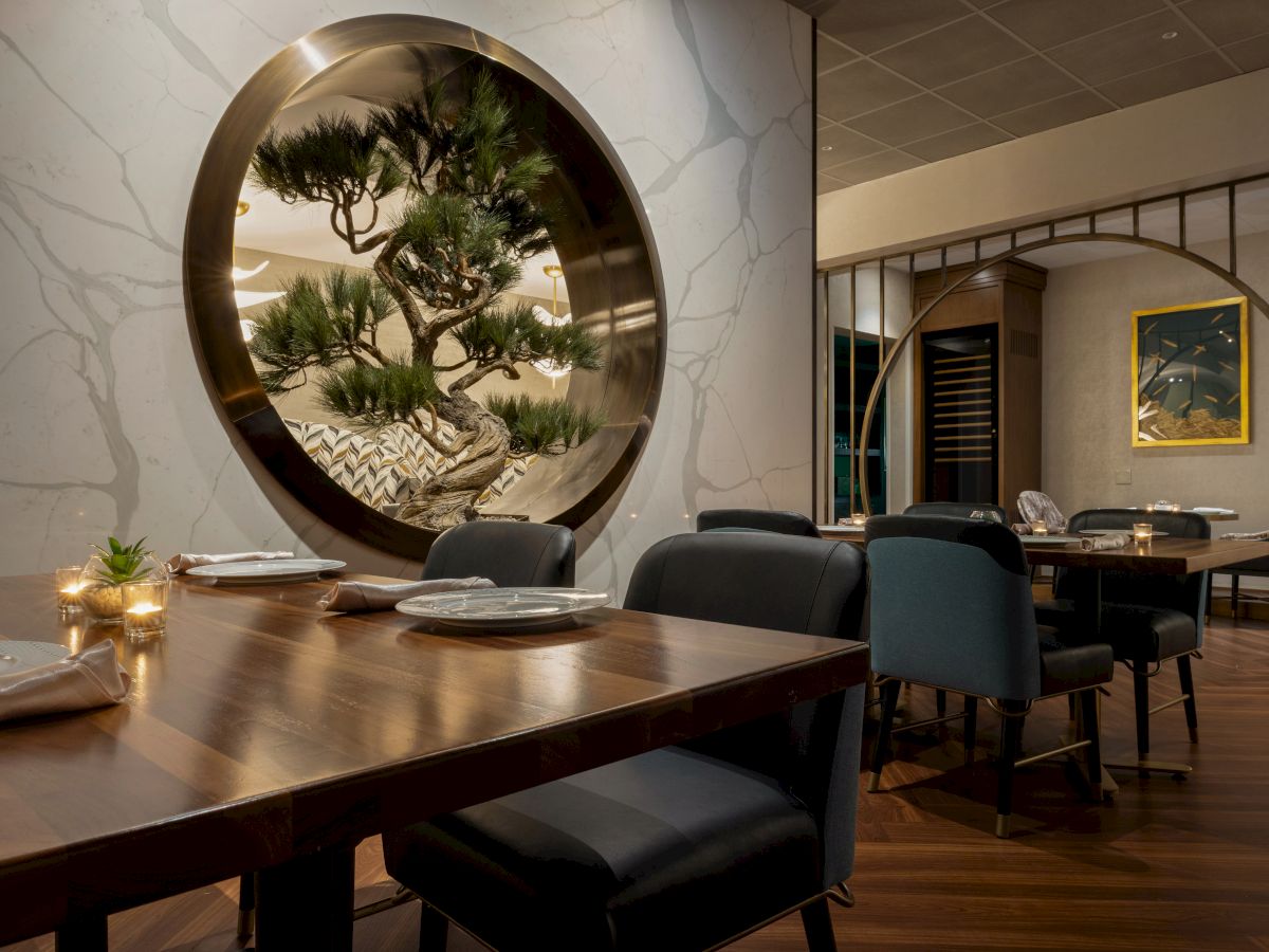 A modern dining room with a round wall window showing a sculpted bonsai-like tree, surrounded by tables, chairs, and wall art.