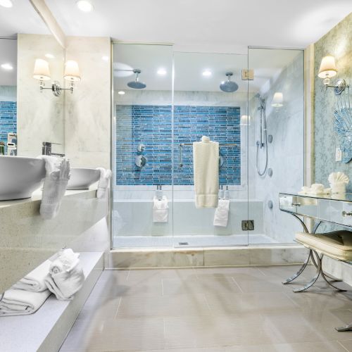 A luxurious bathroom with dual sinks, a large glass shower, and elegant decor. Towels and a bench are neatly arranged.