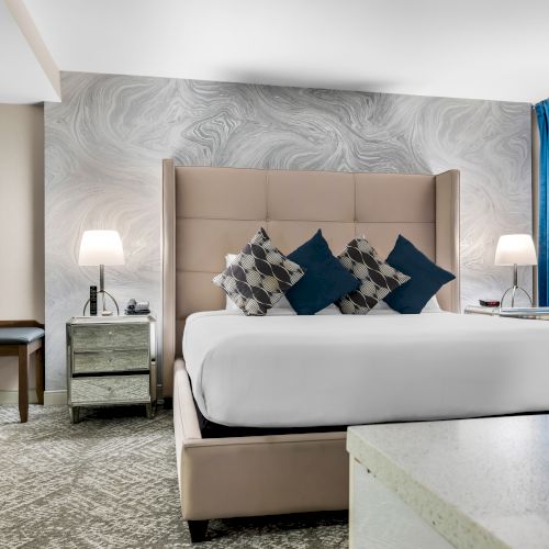 Elegant hotel room with a plush bed, decorative pillows, sleek nightstands with lamps, patterned wallpaper, and rich blue curtains.