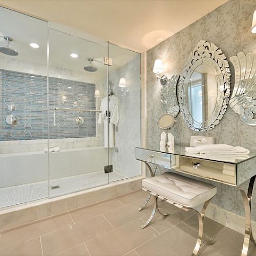 This image shows a luxurious bathroom with a glass-enclosed shower, an ornate mirror, wall sconces, a mirrored vanity table, and elegant wallpaper.