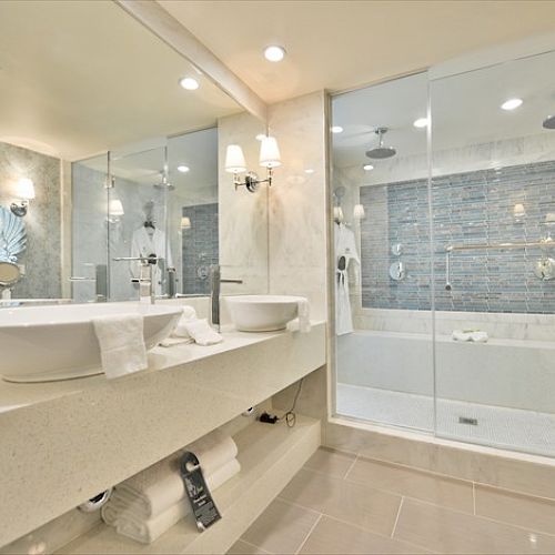 A modern bathroom with a double sink vanity, large mirror, glass-enclosed shower, and white decor, including robes and towels, is shown.