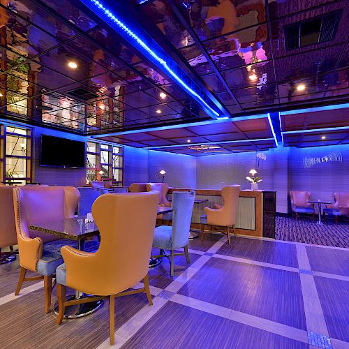 A modern lounge with vibrant blue and purple lighting, featuring cozy seating and tables. The reflective ceiling adds to the ambiance.