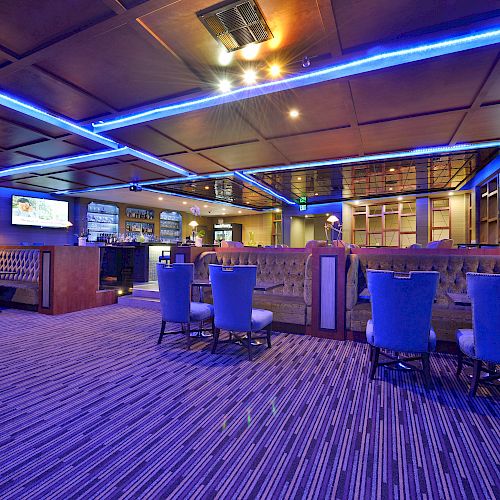 A modern lounge with blue lighting, multiple seating areas, a bar, and televisions on the walls, creating a relaxed and inviting atmosphere.