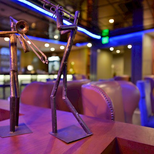 The image shows two metal sculptures resembling musicians in a modern, stylish lounge or bar with blue lighting and comfortable seating.