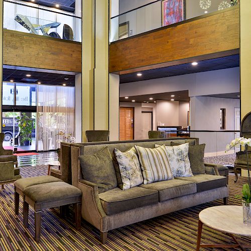 A modern lobby with beige and green furniture, a central couch with pillows, armchairs, round tables, a carpeted floor, and wooden ceilings.