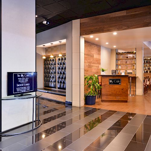 Image shows an entrance to a modern restaurant or café with stylish interior, wooden accents, a counter, plants, and a menu screen.