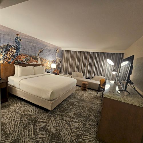 The image shows a modern hotel room with a large bed, a sofa, a desk, a lamp, and patterned carpet, with artwork on the wall.