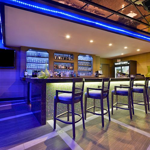 The image shows a modern bar with a counter, bar stools, shelves stocked with bottles, and a TV on the wall, with a blue neon light accent.