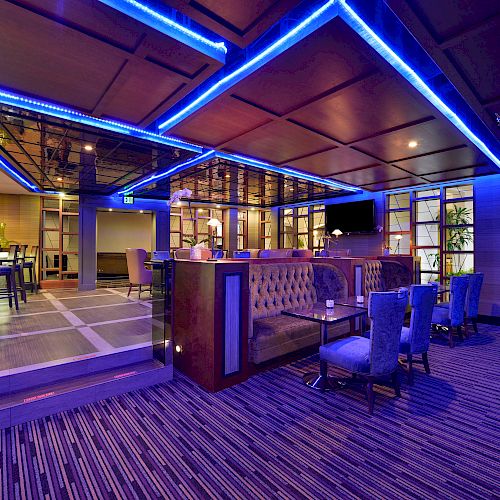 A modern lounge area with blue neon lighting, featuring a bar, high-top stools, cushioned seating, and tables in a stylish, contemporary setting.