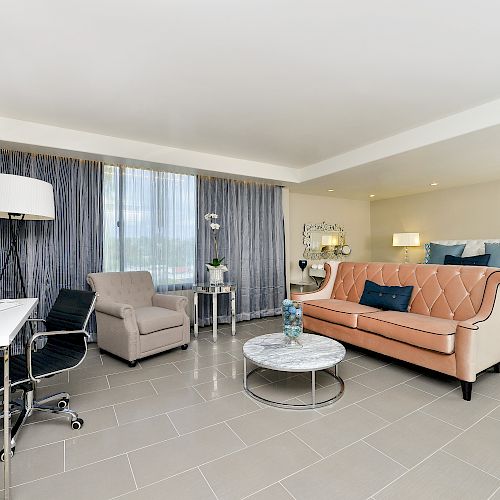 The image shows a modern, spacious hotel room with a bed, sofa, armchair, desk, and various lamps and decor items, featuring a sleek design.