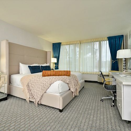 A modern hotel room with a large bed, desk, lamps, and chair. The room is well-lit with natural light from a large window.