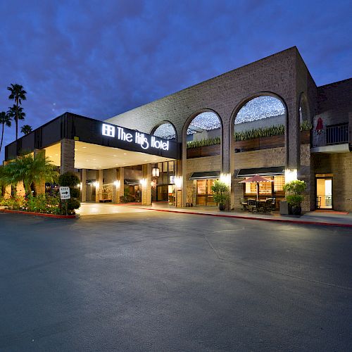 An illuminated hotel entrance with "The Big Hotel" signage, wide driveway, architectural arches, and outdoor plants at dusk.