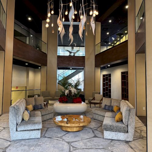 A modern lobby features a curved sofa around a wooden table, with a unique overhead light fixture and decorated plants.