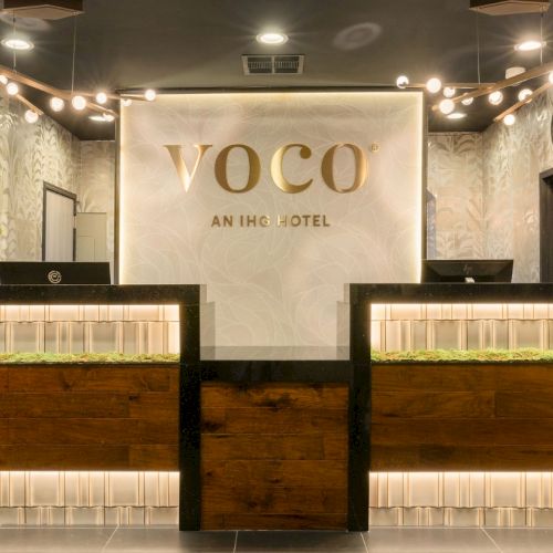 A hotel reception area with a VOCO sign, twin check-in counters, warm lights, and bonsai-style trees.