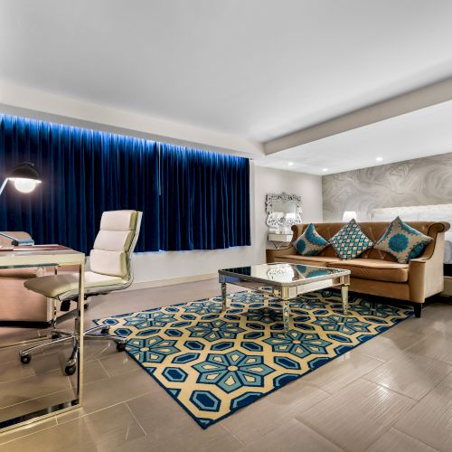 A modern hotel room with a bed, sofa, desk, and large TV. Blue curtains and a patterned rug add color to the bright, spacious area.