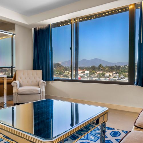 A modern room with a desk, chair, armchair, sofa, mirror coffee table, and large windows showing a scenic mountain view with blue curtains.