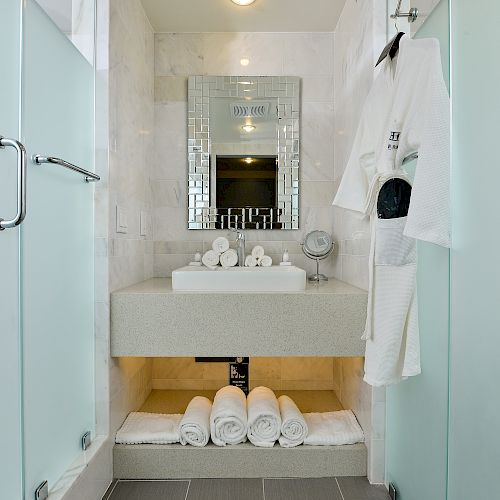 A modern bathroom with frosted glass doors, a sink with towels and toiletries, a wall mirror, and a white bathrobe hanging on the right.