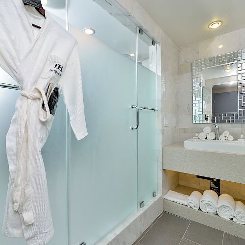 A modern bathroom with a glass shower, white robe, mirror, sink, and folded towels under the counter.