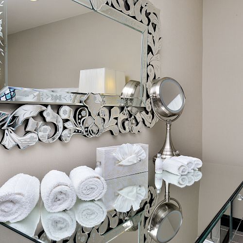 A bathroom vanity with a decorative mirror, rolled white towels, a tissue box, and a small round mirror on a glass counter ending the sentence.