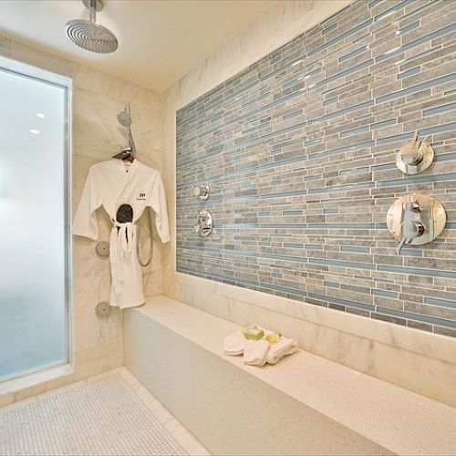 This image shows a luxurious shower room with a glass door, a hanging robe, a towel on a stone bench, and a tiled wall with various faucets.