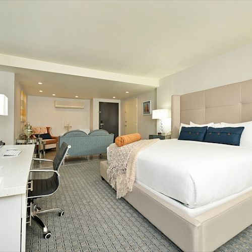 A modern hotel room with a large bed, desk, chair, and a seating area with sofas, all in a neutral color palette, neatly arranged.