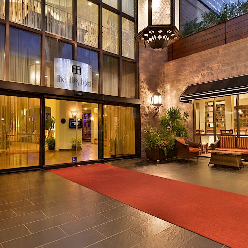 The image shows the entrance of a building with glass doors, a red carpet, outdoor seating, and plants on either side of the entrance door.