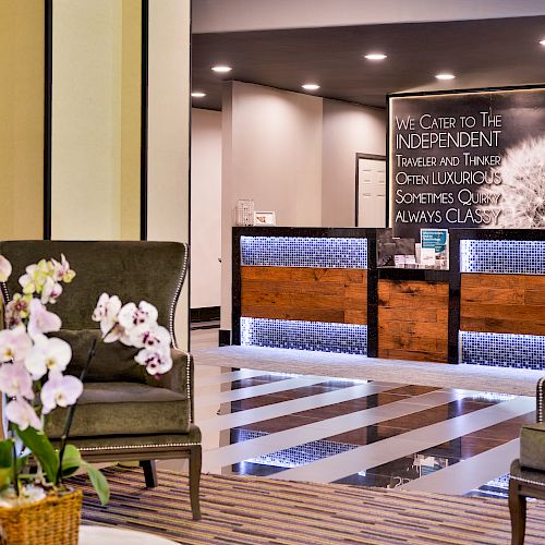 A cozy hotel lobby with two chairs, a flower vase, and a modern reception desk with a sign in the background.