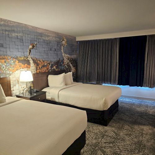 The image shows a hotel room with two beds, a bedside lamp, dark curtains, and a mural on the wall depicting abstract art.