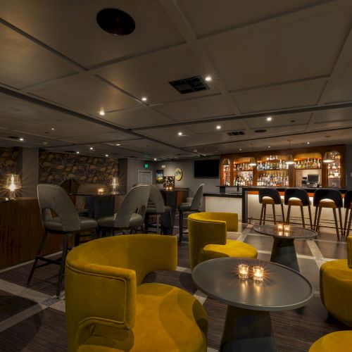 Cozy bar lounge with booths, yellow seating, a patterned wall, and a backlit liquor shelf behind the counter.